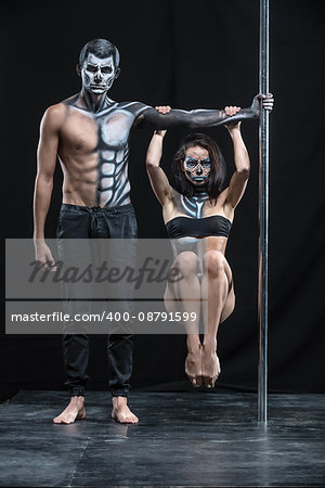 Perfect couple of pole dancers in the dark studio. Man holds a pylon with the left hand. Girl hangs with tighten knees on the guy's left arm. They wear black sportswear and have a horrific body-art.