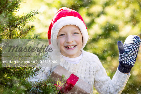 beautiful smiling boy in santa's hat, sweater and mittens holding nicely wrapped christmas gift by the tree or in the forest enjoying snowy cold winter weather, holiday or happiness concept
