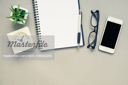Top view of office desk table with open spiral notebook, white alarm clock and mobile phone on grey background with vintage filter