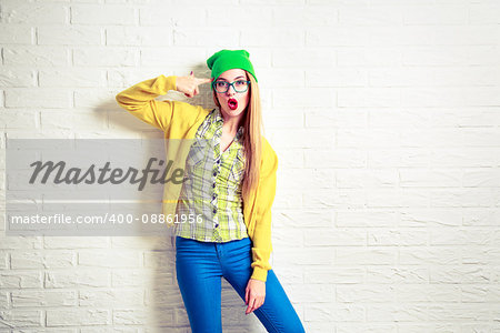 Funny Hipster Girl at White Brick Wall Background. Street Syle Teenage Going Crazy. Circling a First Finger by Her Temple. Trendy Fashion Outfit in Spring or Autumn. Copy Space.