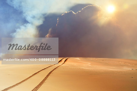 Vehicle tracks through desert and dunes leading into a sand, smoke and cloud filled sky. Digital photo manipulation.