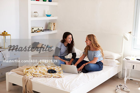 Two girls sitting on bed using laptop looking at each other