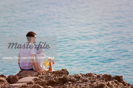family of two, father and son, sitting on the rocks enjoying sunset at the tropical island