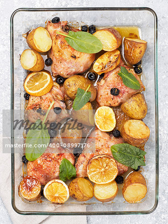 Top view of chicken thighs with potatoes, lemon and black olives, cooked in oven on gray concrete background. Baked chicken leg quarter in heat-proof glass. Vertical.