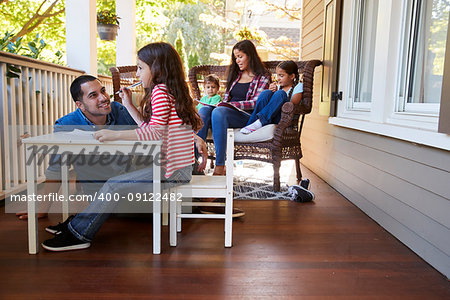 Family Sit On Porch Of House Reading Books And Playing Games