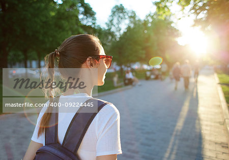 back view of a young attractive woman in white t-shirt with small city backpack at sunset on the walkway in park