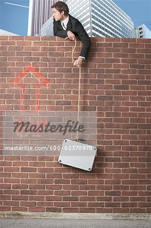 Businessman Leaning over Brick Wall, holding onto Rope with Briefcase Attached