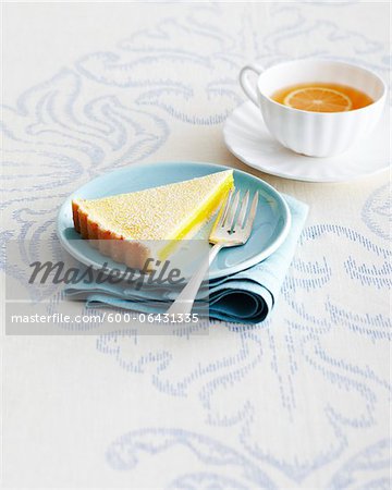 Slice of Lemon Tart and Fork on Blue Plate with Cup and Saucer of Herbal Tea on Tablecloth in Studio