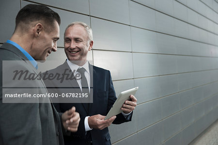 Mature businessmen standing in front of wall, talking and looking at tablet computer