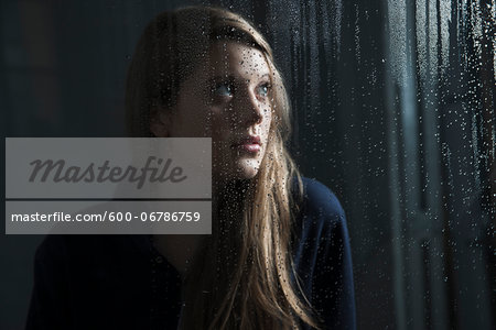 Portrait of young woman behind window, wet with raindrops, looking up