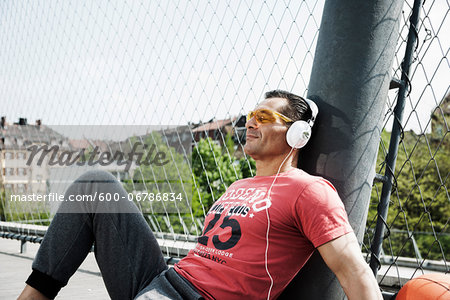 Mature man sitting on outdoor basketball court wearing headphones and listening to music, Germany