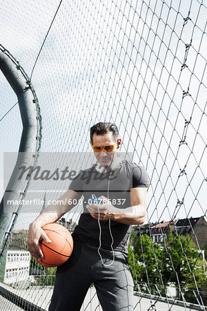 Mature man standing on outdoor basketball court holding basketball and looking at MP3 player, Germany