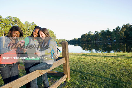 Pre-teen girls sitting on fence, looking at tablet computer and cellphone, outdoors