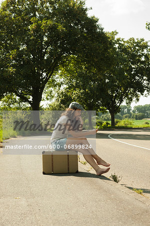 Teenaged girl sitting on suitcase on the side of the road, looking at cell phone, in summer, Germany