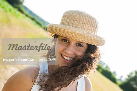 Close-up portrait of teenaged girl standing in field, wearing straw hat, smiling at camera, Germany