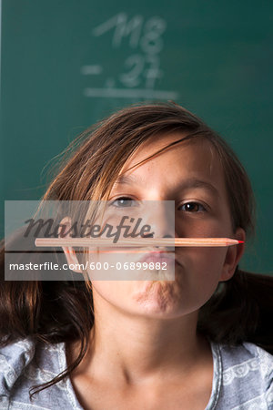 Portrait of girl standing in front of blackboard in classroom, holding pencil with mouth, Germany