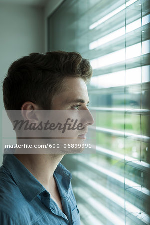 Close-up portrait of young man, looking out window through blinds, Germany