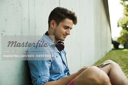 Young man sitting next to wall of building outdoors, with headphones around neck and looking at cell phone, Germany