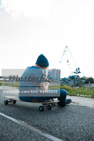 Backview of boy sitting on skateboard outdoors in an industrial area and looking at cell phone, Germany