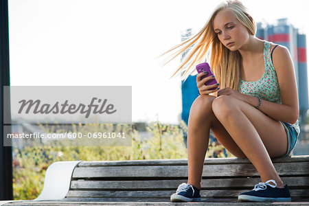 Teenage girl sitting on bench outdoors, looking at cell phone, Germany