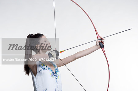 Side view of young, woman archer, aiming bow and arrow, studio shot on white background