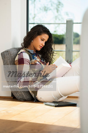 Teenage girl sitting on floor next to window and reading book, Germany
