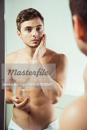 Young man looking in bathroom mirror, applying cologne to face, studio shot