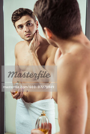 Young man looking in bathroom mirror with towel wrapped around waist, applying cologne to face, studio shot