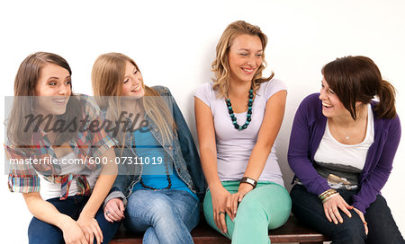 Four, young women sitting on bench together, laughing and looking at each other, studio shot on white background
