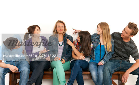 Six, young men and women sitting on bench together, laughing and looking at each other, studio shot on white background