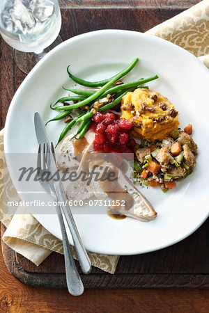 Overhead image of a holiday dinner plate containing turkey breast, gravy, stuffing, sweet potato, green beans, cranberries, walnuts and fresh rosemary. Cutlery, ice water, and a napkin are also present on a wooden cutting board on a table top.