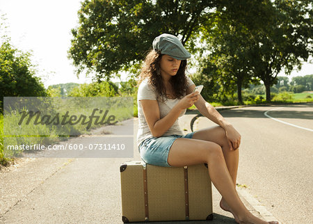 Teenage girl sitting on suitcase on the side of the road, looking at cell phone in summer, Germany