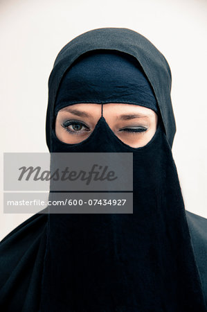 Close-up portrait of young woman wearing black, muslim hijab and muslim dress, winking and looking at camera, eyes showing eye makeup, studio shot on white background