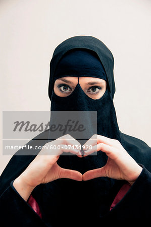 Close-up portrait of young woman wearing black, muslim hijab and muslim dress, making heart shape with hands, looking at camera, eyes showing eye makeup, studio shot on white background Close-up portrait of young woman wearing black, muslim hijab and muslim dress, making heart shape with hands, looking at camera, eyes showing eye makeup, studio shot on white background
