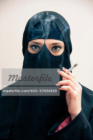 Close-up portrait of young woman wearing black, muslim hijab and muslim dress, holding cigarette and smoking, looking at camera, eyes showing eye makeup, studio shot on white background