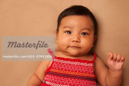 Close-up portrait of Asian baby lying on back, wearing red dress, looking at camera and smiling, studio shot on brown background
