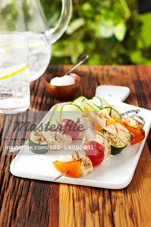 BBQ Chicken and vegetable skewers on a white serving platter with lemon sparkling water on a wood picnic table, studio shot