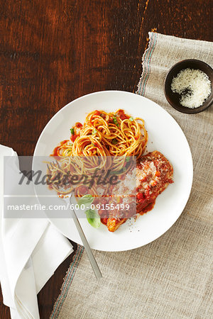 Plate of chicken parmesan with spaghetti and tomato sauce and a side dish of cheese with a fork and napkin on a wooden background