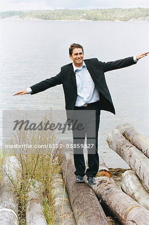 Businessman balancing on logs beside sea with arms out, smiling