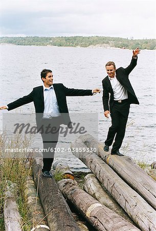 Two businessmen balancing on logs beside sea with arms out, smiling