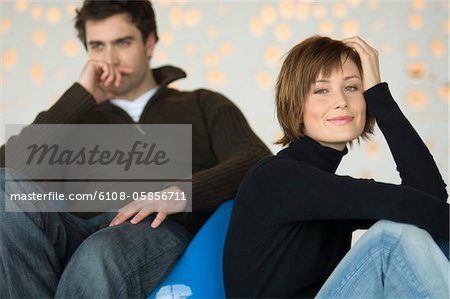Couple sitting in living-room, woman looking at the camera, man thinking in the background