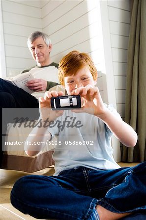 Boy sitting cross-legged, taking picture with camera phone, senior man reading in background