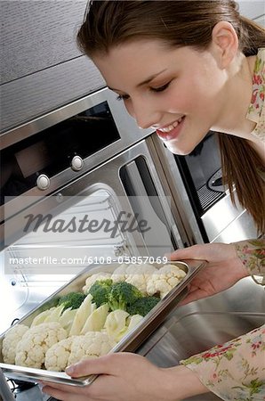 Young smiling woman putting broccolis and cauliflowers into an oven
