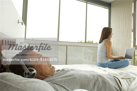 Couple in bedroom, man listening to music, woman using laptop, indoors