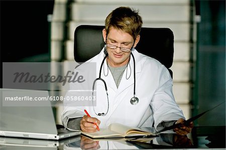 Male doctor sitting at a desk in his office and writing in a notebook
