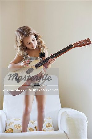 Cute little girl standing on a sofa and playing a guitar