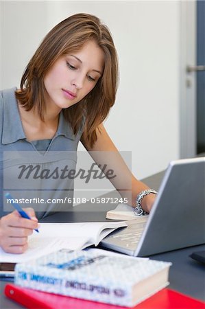 Beautiful woman writing in book while using laptop in classroom