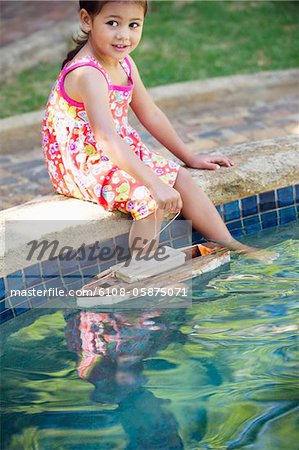 Girl sitting at edge of swimming pool with toy boat in water