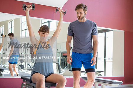 Trainer looking at a young woman lifting dumbbells in a gym