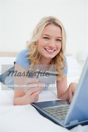 A close up shot of a woman lying on a bed ordering items online with her credit card and laptop.